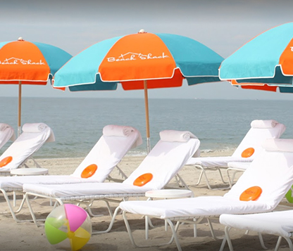 Several Frankford Emerald Coast umbrellas with orange and turquoise fabric shading white beach lounge chairs by the shore